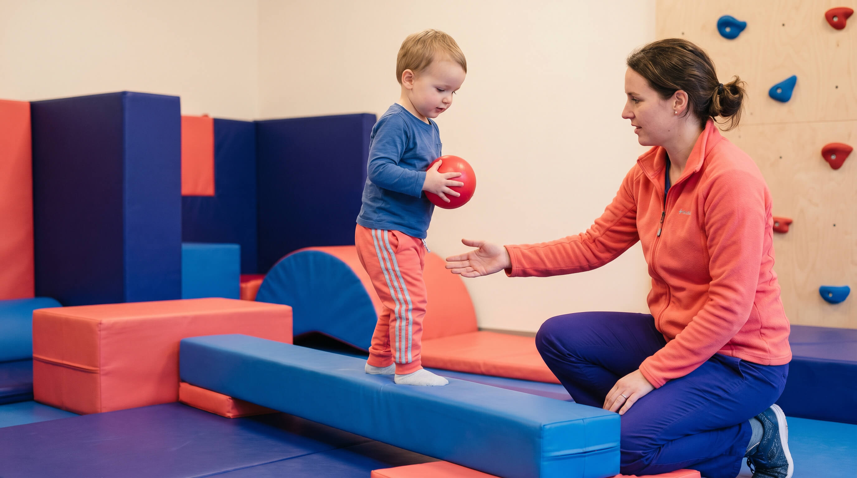 Kind macht Balance Übung mit Ball in seinen Händen. Unterstützt durch eine Therapeutin